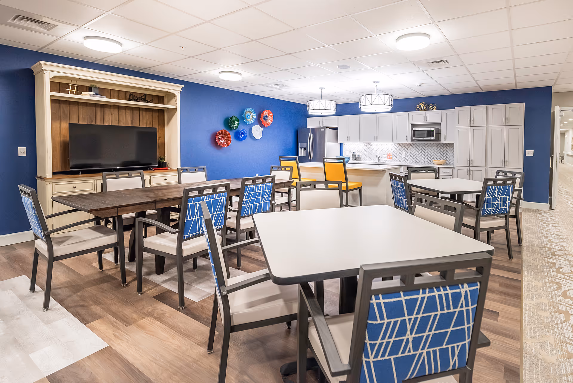 A bright and modern common area with multiple tables and chairs, a large TV on a wooden entertainment unit, and a kitchen area with white cabinets, a refrigerator, microwave, and an island with yellow bar stools. The walls are painted blue and decorated with colorful wall art.