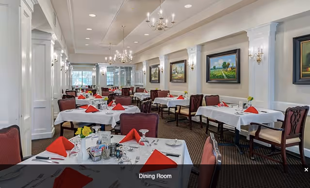 A spacious dining room with multiple tables covered in white tablecloths, each set with red folded napkins, glassware, and silverware. The room features elegant chandeliers, wall sconces, framed landscape paintings, and large windows allowing natural light to enter.