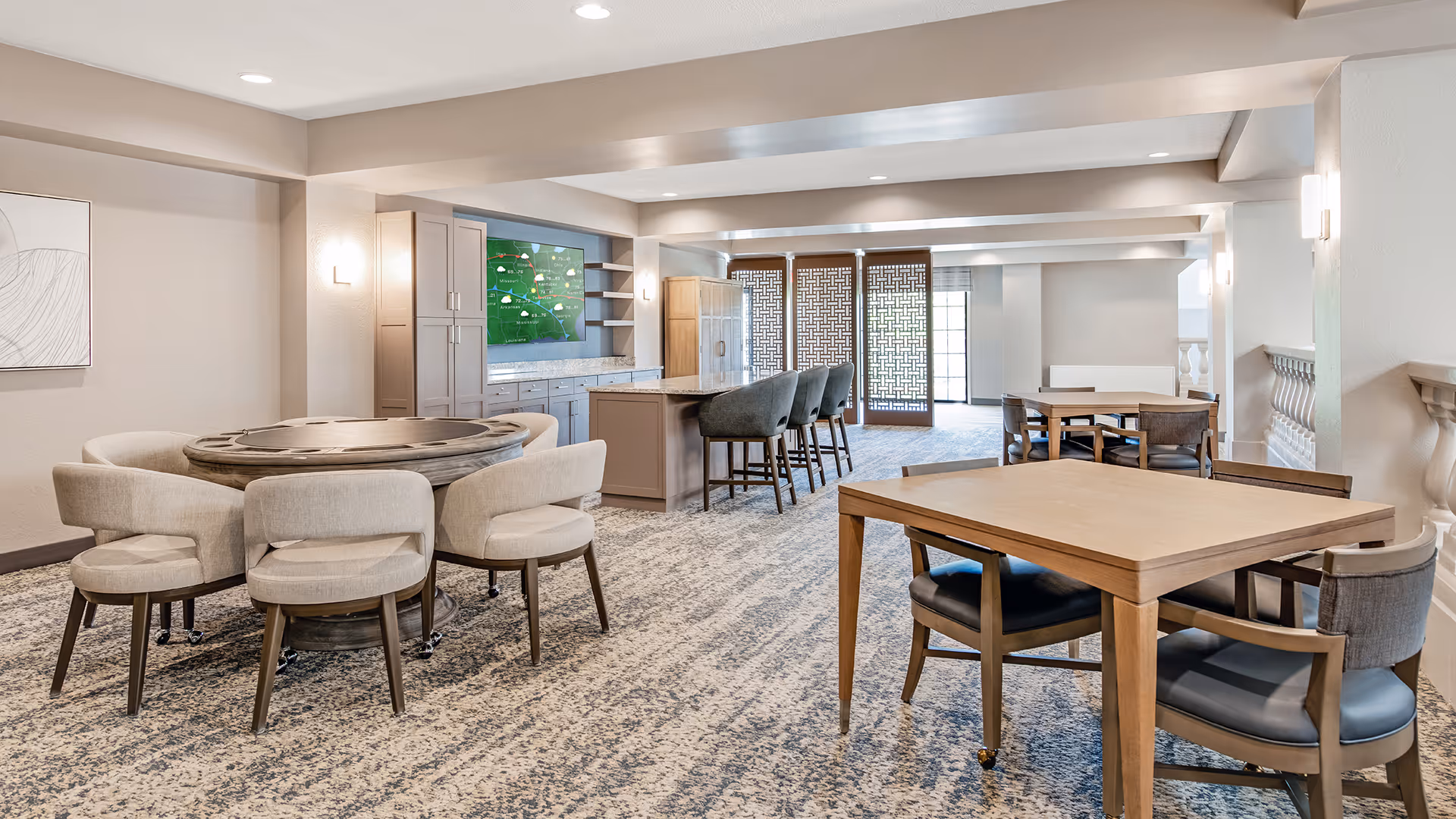 A spacious, well-lit common area with multiple seating arrangements including a round table with cushioned chairs and square wooden tables with chairs. The room features a carpeted floor, neutral-colored walls, built-in cabinetry with a large TV screen, and decorative wooden panels near the windows allowing natural light to enter.
