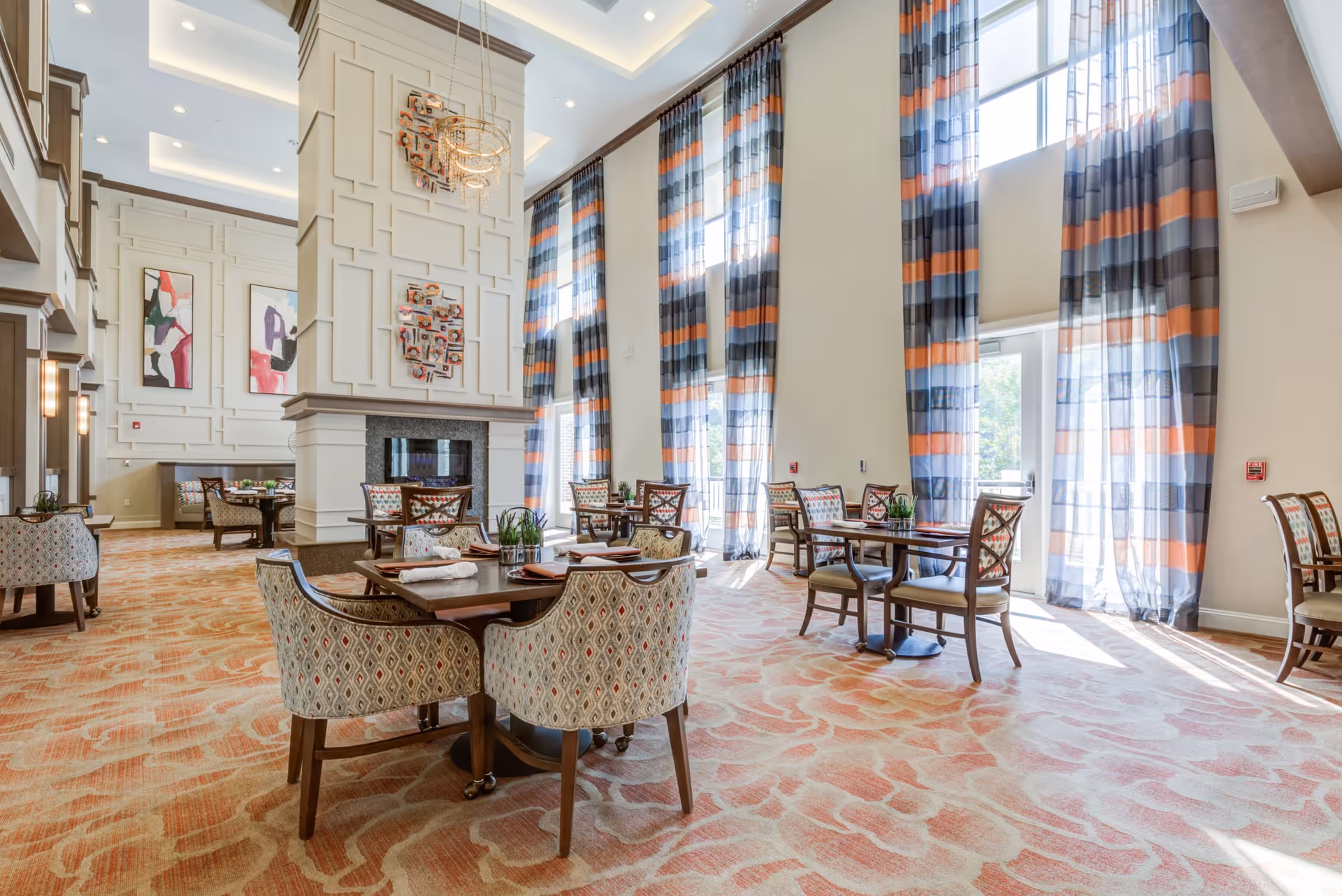 A spacious and well-lit dining room with multiple tables and chairs arranged neatly. The room features tall windows with colorful striped curtains in shades of blue, orange, and gray, allowing natural light to fill the space. The walls are decorated with modern art pieces and a large, elegant chandelier hangs from the ceiling. The carpet has a patterned design in warm tones, complementing the overall decor.