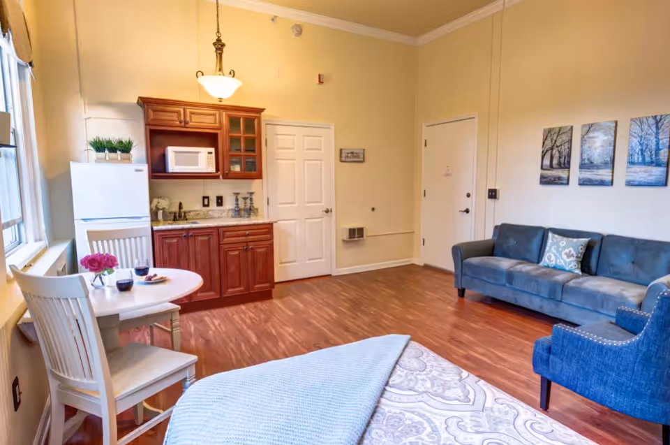 A cozy living space featuring a small kitchenette with wooden cabinets, a microwave, and a refrigerator. There is a round dining table with two chairs near a window, set with two glasses of red wine and a small vase with pink flowers. The room also includes a blue sofa, a blue armchair, and a bed with a patterned bedspread and a folded blanket. The walls are light-colored with three framed pictures of trees hanging above the sofa. The floor is wooden, and the room is well-lit with natural light from the window and a ceiling light fixture.