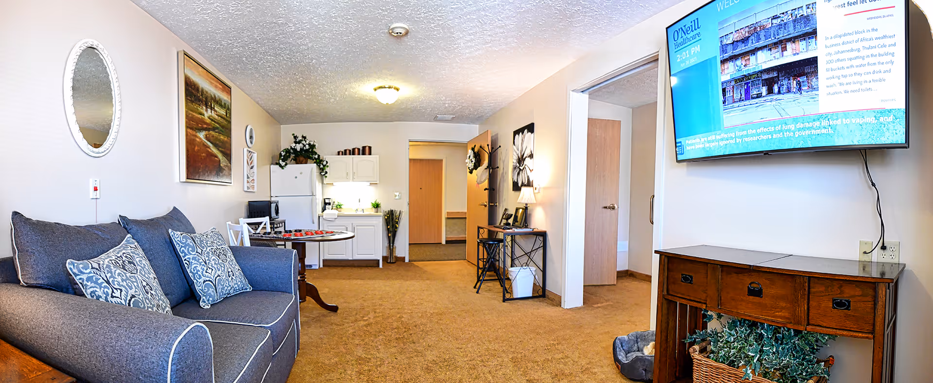 Bright senior living room with a couch, kitchenette, mounted TV, small dining table and open doorways.