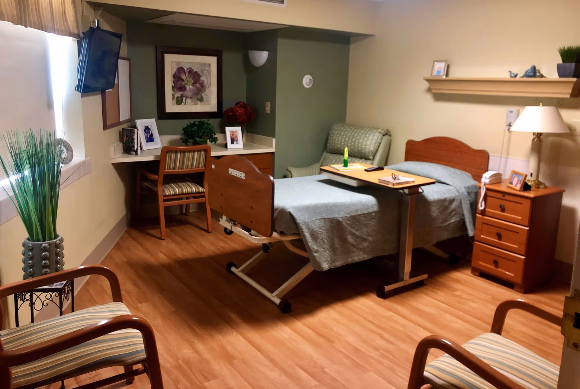 A well-lit room in a healthcare facility featuring a hospital bed with a green bedspread, an overbed table with items on it, a wooden nightstand with a lamp and telephone, a green upholstered chair, a desk with a wooden chair, framed pictures, a wall-mounted TV, and decorative plants.