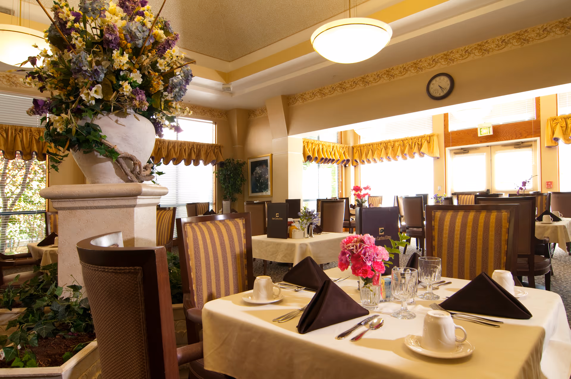 A bright and elegant dining room with tables set for a meal, featuring white tablecloths, folded black napkins, glassware, and floral centerpieces. The room has large windows with yellow valances, striped upholstered chairs, and a large decorative flower arrangement on a pedestal in the center.