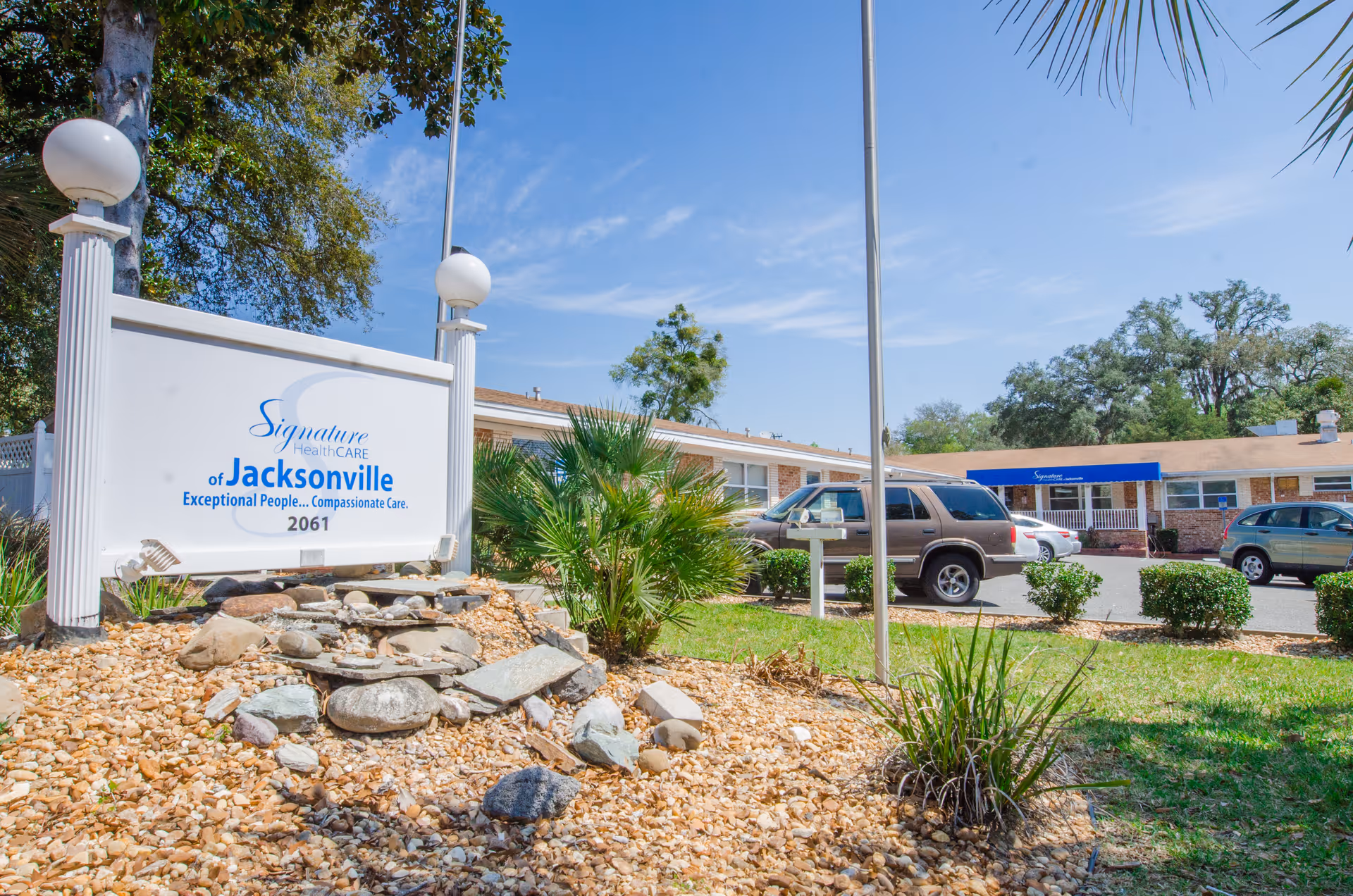 Outdoor view of the entrance area of a senior living facility with a white sign reading 'Signature HealthCARE of Jacksonville Exceptional People... Compassionate Care. 2061' surrounded by rocks and plants. In the background, there is a single-story building with a blue awning and several parked cars under a clear blue sky.