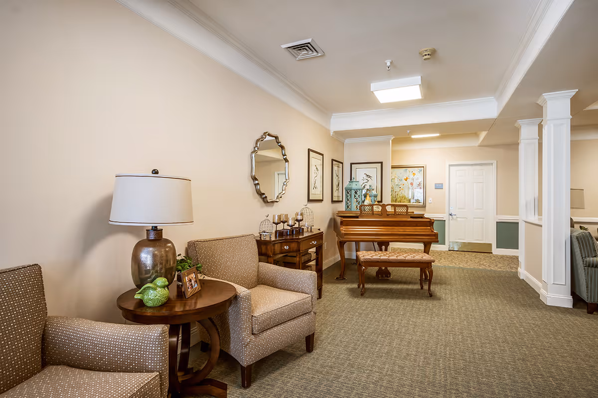 A cozy interior common area with beige walls and carpeted floor featuring two patterned armchairs around a small wooden side table with a lamp and decorative items. A wooden console table with candles and birdcages sits against the wall beneath a decorative mirror and framed artwork. A wooden piano with a cushioned bench is positioned near a white door, with additional framed artwork on the walls.