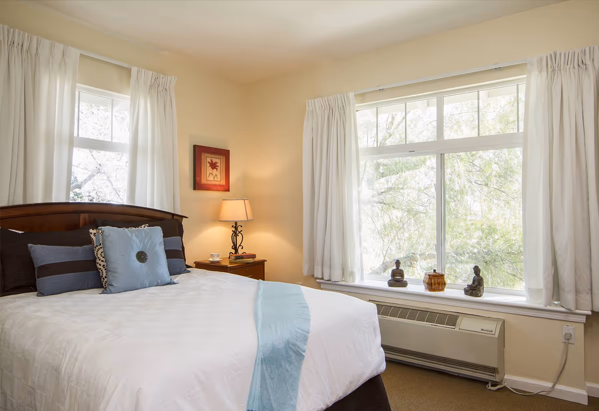 A cozy bedroom with a neatly made bed featuring white and blue bedding. The room has two large windows with white curtains, letting in natural light and showing green trees outside. A wooden nightstand with a lamp, a cup, and a book is beside the bed. Decorative items including two small statues and a container are placed on the windowsill.