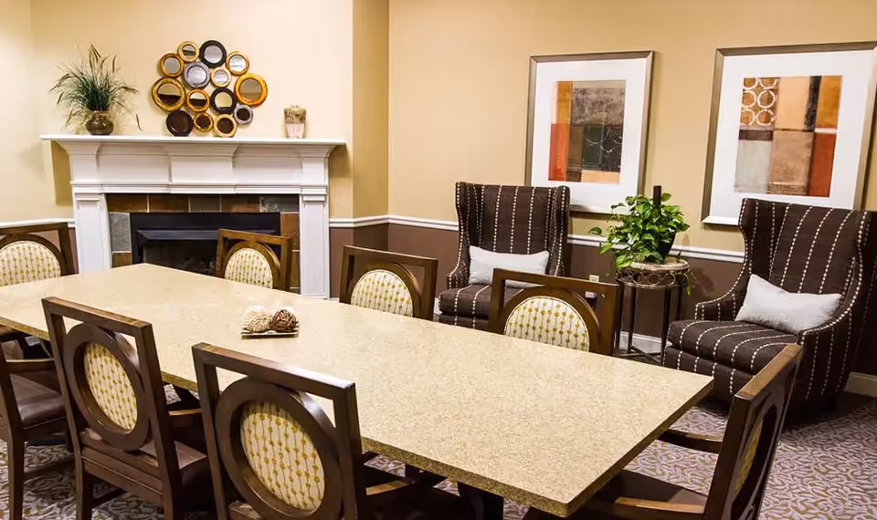 A dining room with a long rectangular table surrounded by wooden chairs, upholstered armchairs, a fireplace mantel, and framed wall art.
