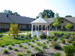 A landscaped garden area with a white wooden gazebo in the center, surrounded by various shrubs and small trees. The background shows a single-story building with a dark roof and beige walls under a clear blue sky.