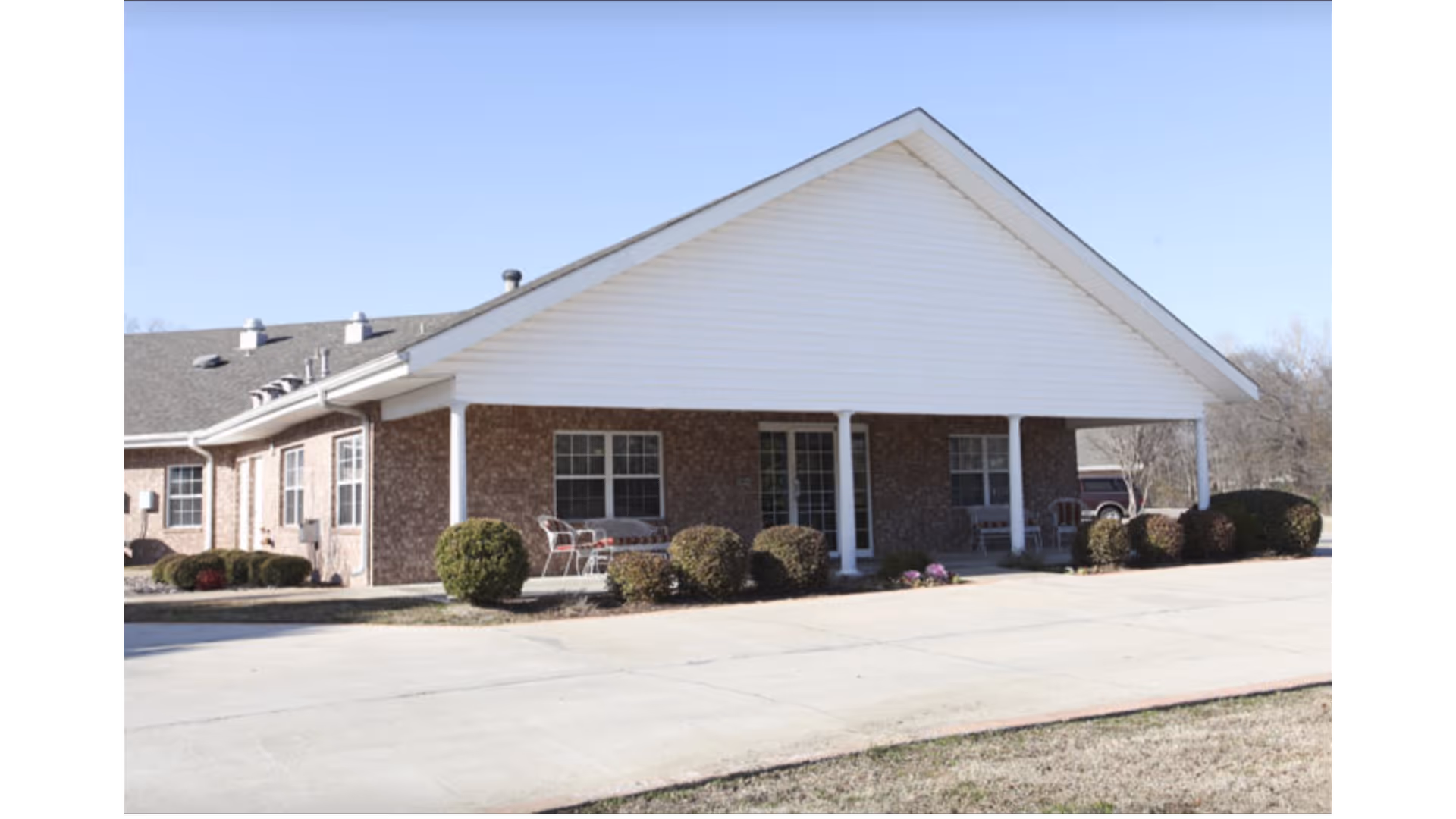 Exterior view of a single-story brick building with a white gabled roof and a covered porch supported by white columns. The porch has outdoor chairs and small bushes planted along the front. The sky is clear and blue.