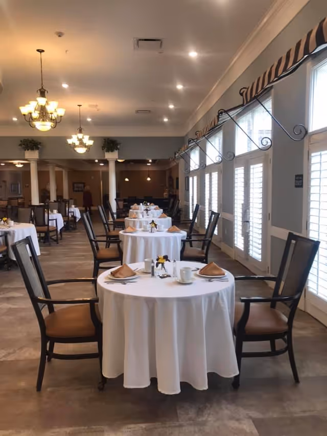 A dining room with round tables covered in white tablecloths, each set with folded napkins, cups, and small flower arrangements. The room has wooden chairs with armrests, large windows with white shutters, and decorative valances. Ceiling lights and chandeliers illuminate the space.