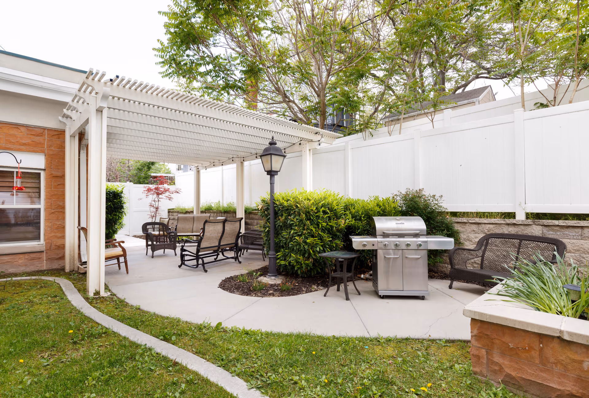 Outdoor patio area with a white pergola covering seating that includes cushioned chairs and a bench. There is a stainless steel grill, a small table, a lamp post, and greenery including bushes and trees surrounding the space. A white fence encloses the area.