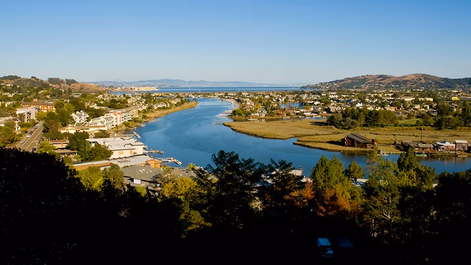 A scenic view of a river winding through a residential area with houses, trees, and hills in the background under a clear blue sky.