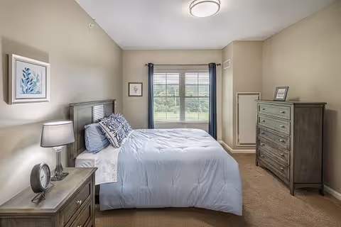 A neatly made bed with light blue bedding and decorative pillows in a bedroom. The room has beige walls, a window with dark curtains, a wooden nightstand with a lamp and clock, a wooden dresser with a framed picture on top, and carpeted flooring.