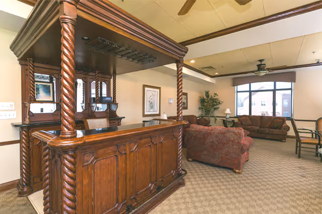A furnished common room with a carved wooden bar, upholstered sofas, a coffee table, and a large window.