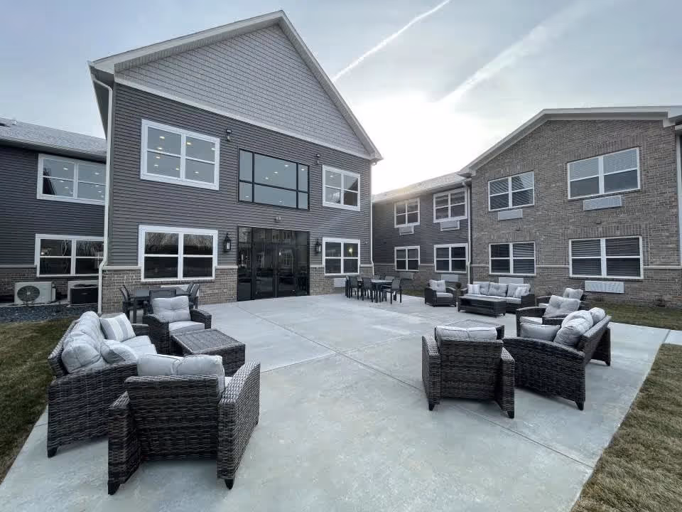 Outdoor patio area at Autumn Trace Bedford Assisted Living featuring multiple sets of cushioned wicker chairs and sofas arranged on a concrete surface, surrounded by two-story residential-style buildings with multiple windows under a partly cloudy sky.