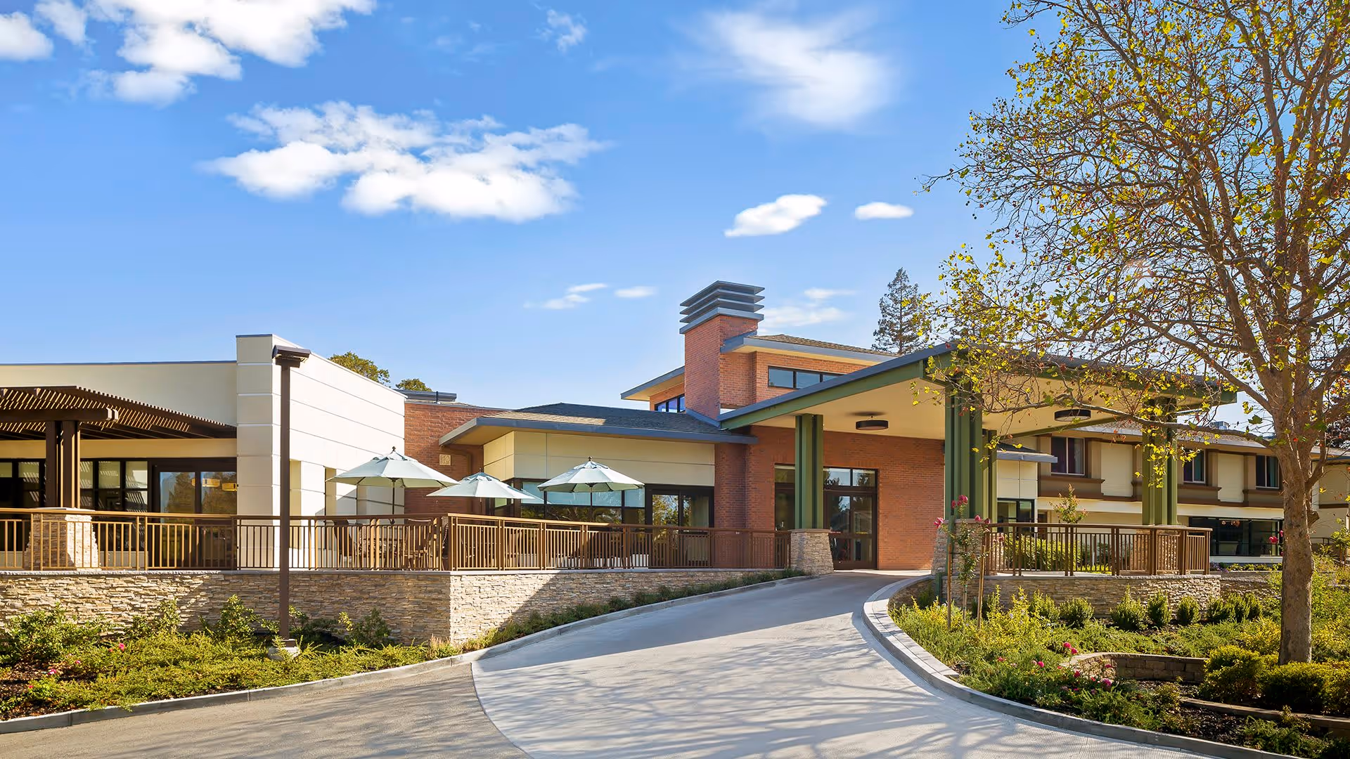 Exterior view of Atria Walnut Creek senior living facility on a sunny day, showing a modern building with a covered entrance, outdoor patio with umbrellas, and landscaped greenery.
