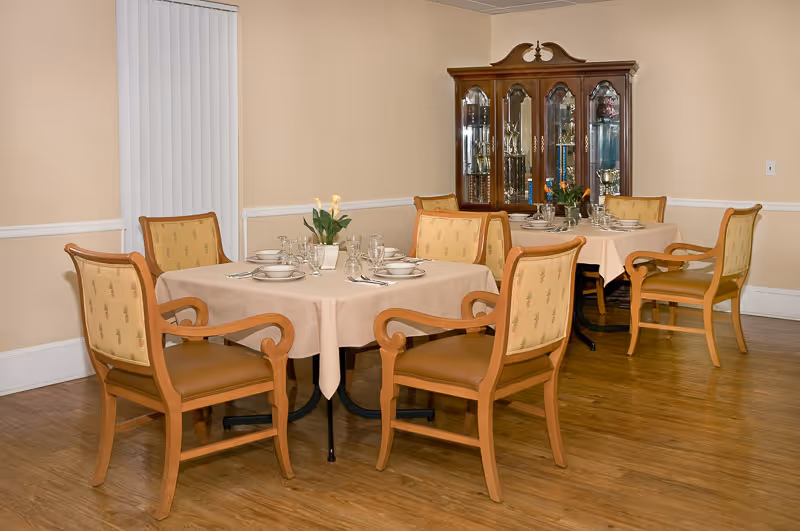 A dining room with two tables covered with beige tablecloths, each surrounded by four wooden chairs with cushioned seats and backs. The tables are set with plates, glasses, and silverware, and each has a small floral centerpiece. In the background, there is a wooden china cabinet displaying trophies and decorative items. The room has light-colored walls and wooden flooring.