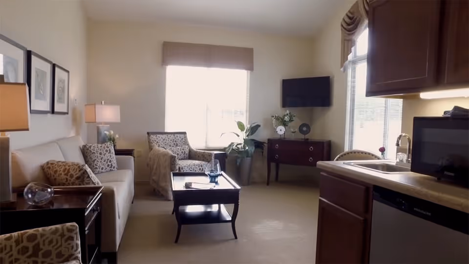 A cozy living room area in a senior living facility featuring a beige sofa with patterned cushions, a matching armchair, a wooden coffee table with a glass and a book on it, a side table with a lamp, a flat-screen TV mounted on the wall, and a kitchenette with a sink, microwave, and dishwasher. Large windows allow natural light to brighten the room.