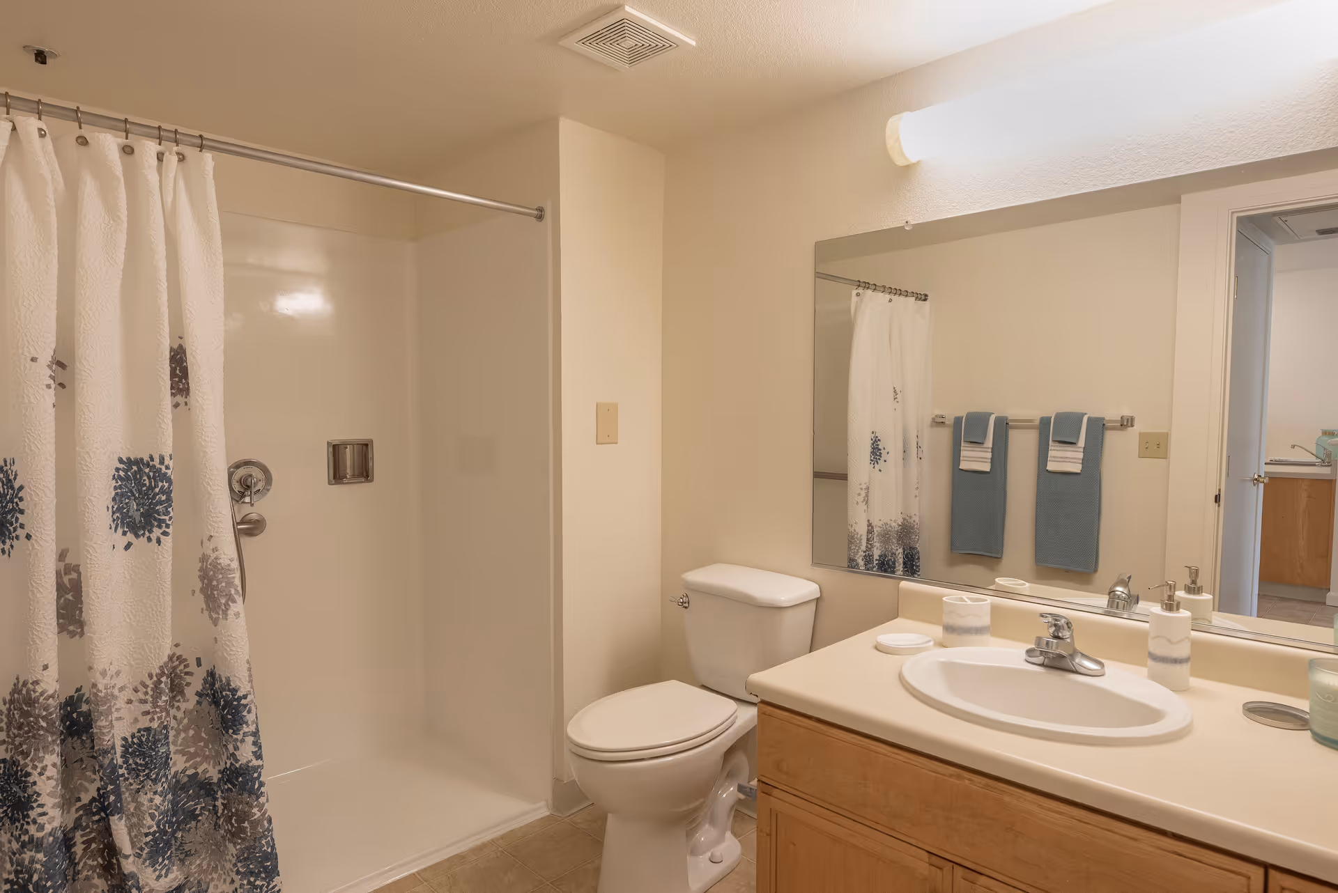 A clean bathroom with a white shower area and a shower curtain with blue and gray floral patterns. There is a white toilet next to a wooden vanity with a white sink and countertop. Above the sink is a large mirror reflecting two blue towels hanging on a towel rack and part of an adjacent room.
