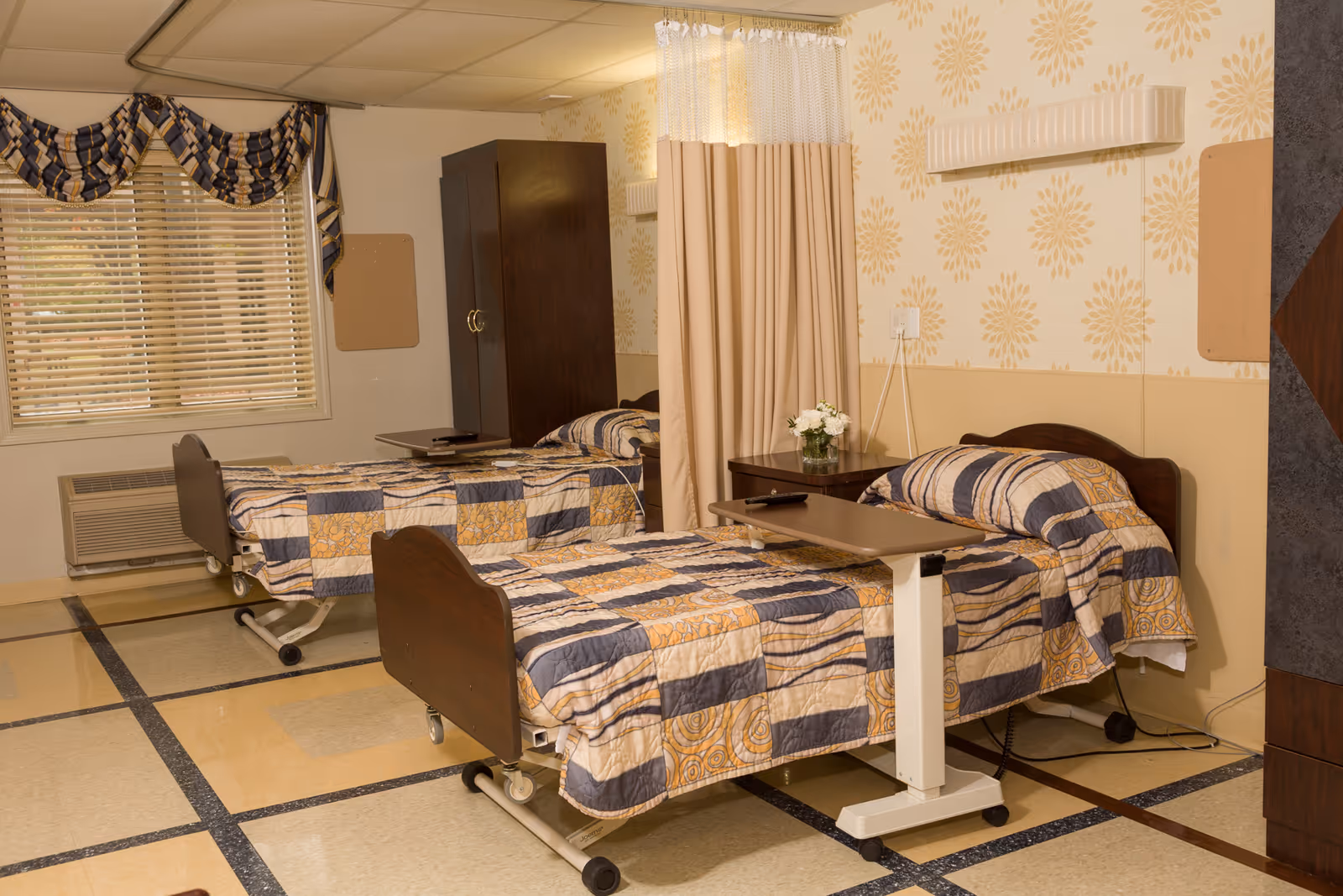 A shared bedroom in a care facility with two single beds, each covered with a patterned quilt in shades of blue, beige, and yellow. Each bed has an adjustable overbed table. There is a wooden nightstand with a small vase of white flowers between the beds. The room has a window with blinds and a decorative valance, a dark wooden wardrobe, and a privacy curtain. The walls are decorated with a light floral pattern and the floor has a tiled design.