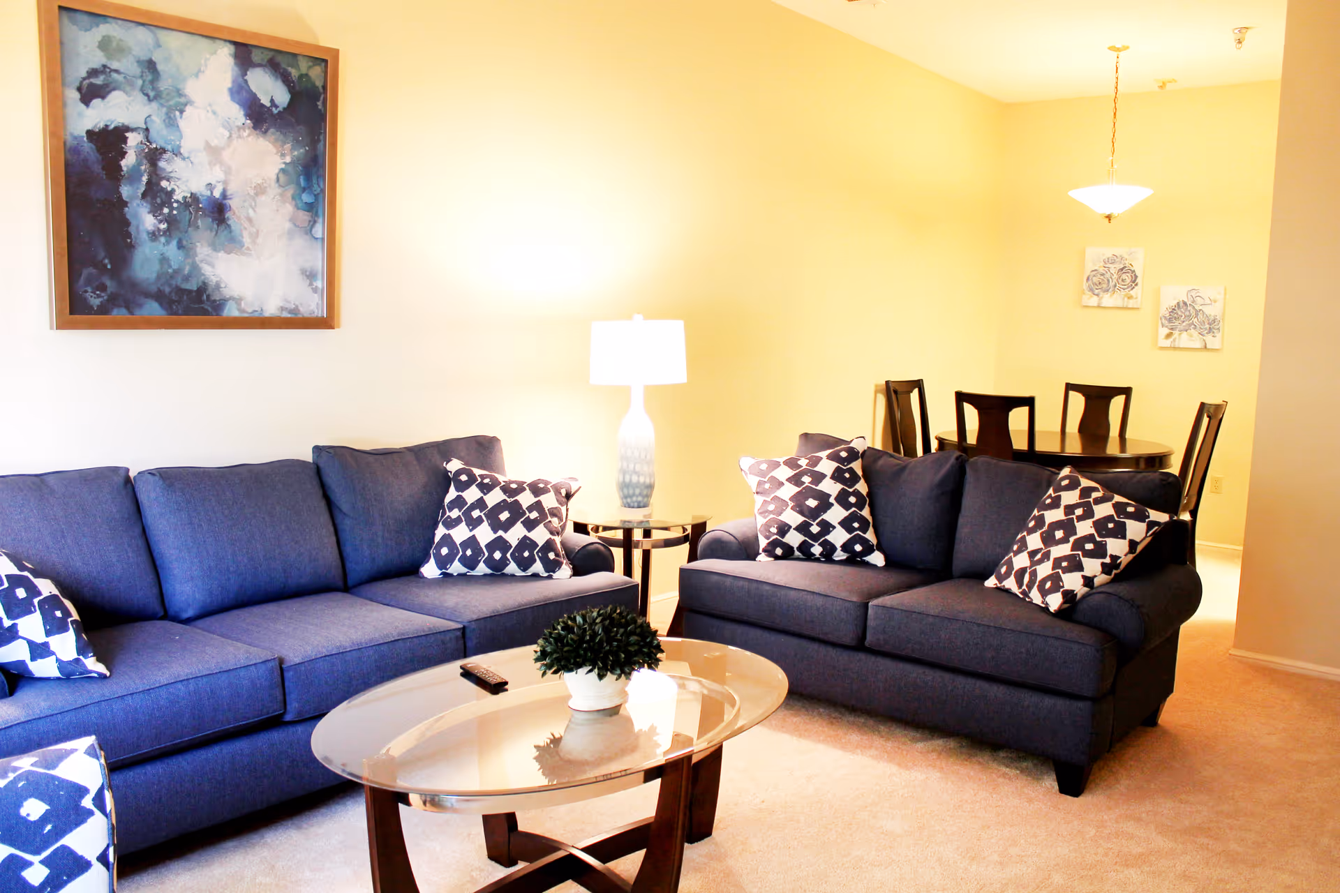 Bright living room with two navy sofas, patterned throw pillows, a glass coffee table, lamp, and a dining area in the background.