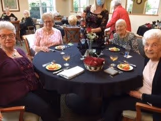 Four elderly women sitting around a round table with black tablecloth in a dining area, each with a plate of food and glasses of water. Other people are visible in the background, and the room has large windows letting in natural light.