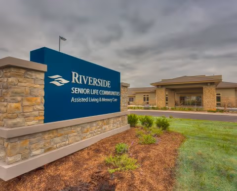 Exterior view of the entrance sign for Riverside Senior Life Communities, featuring a large blue sign with white text and logo, set against a landscaped area with grass and small plants, with the building visible in the background under a cloudy sky.