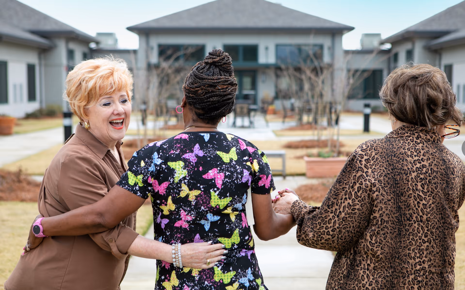 Three women standing outside in a courtyard area of a senior living facility. One woman with short blonde hair and a brown shirt is smiling and holding the arm of a woman in colorful butterfly print scrubs, who is holding hands with another woman wearing a leopard print jacket. The background shows buildings and a paved walkway with some landscaping.