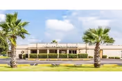 Front exterior view of Valley Grande Manor building with a beige facade, a row of windows, and two palm trees in the foreground under a partly cloudy sky.