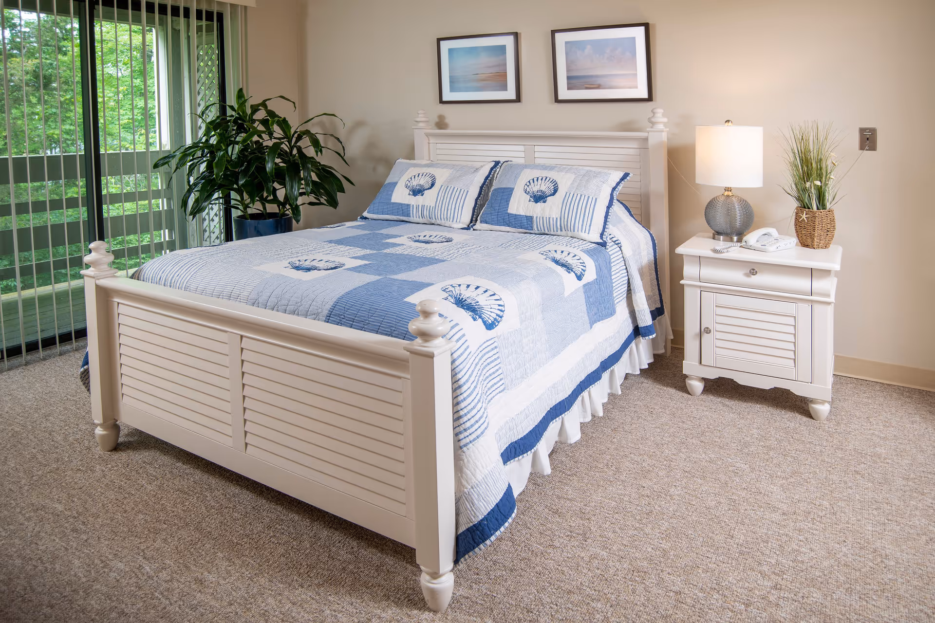 A neatly made bed with a white wooden frame and a blue and white quilt featuring seashell patterns. There are two framed pictures hanging on the beige wall above the bed. To the right of the bed is a white nightstand with a lamp, a telephone, and a small plant. A large green potted plant is placed near a sliding glass door with vertical blinds, allowing natural light to enter the room.