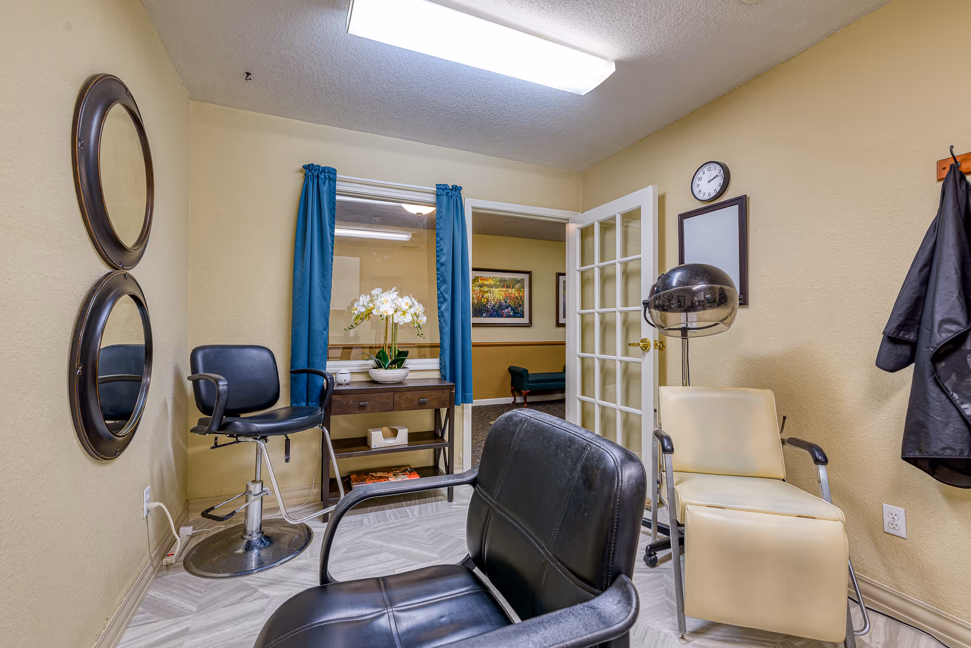 Interior view of a small salon area in a senior living facility with two salon chairs, a hair dryer, two round mirrors on the wall, a small table with a flower arrangement, and a glass-paneled door leading to another room.