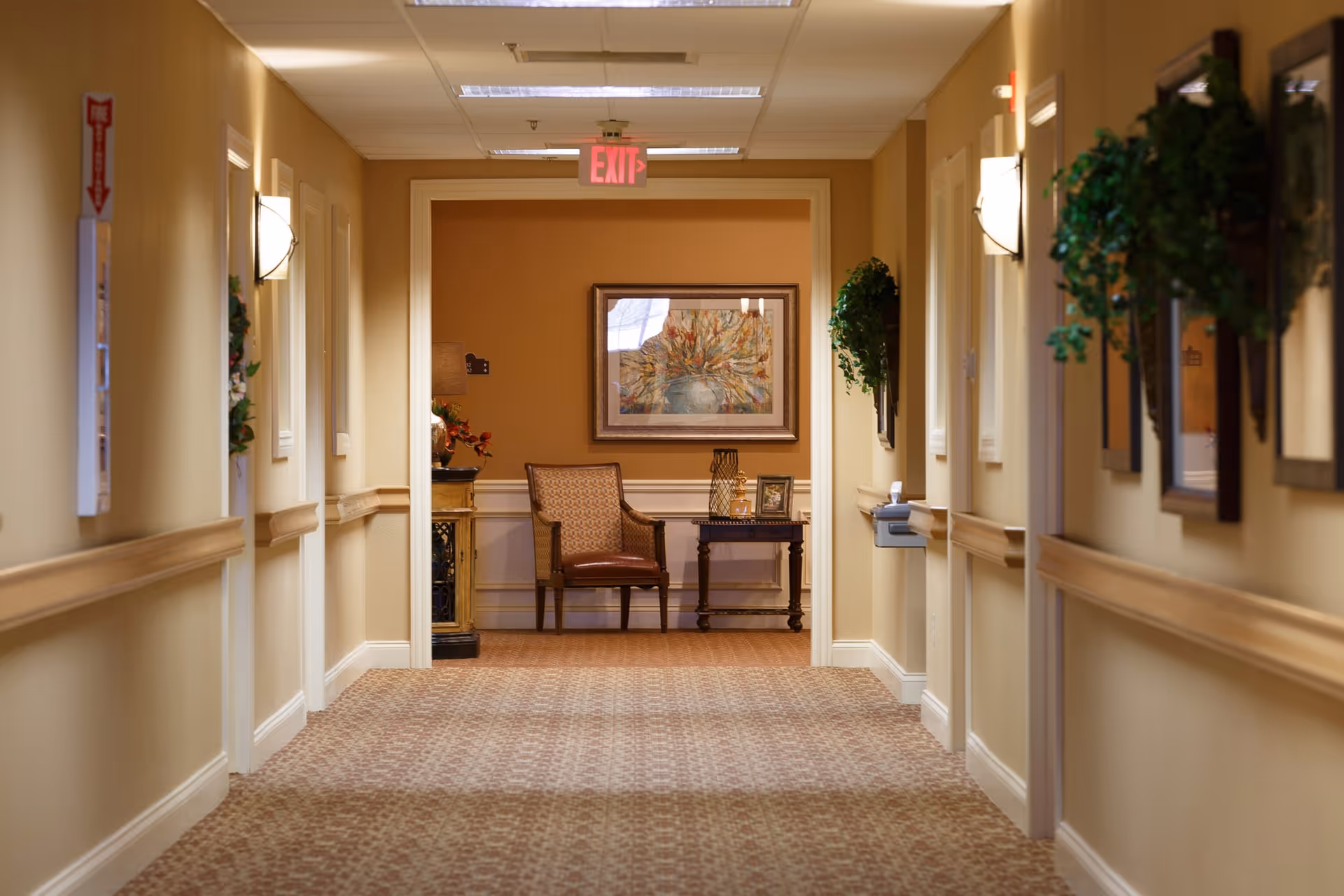A well-lit hallway in a senior living facility with beige walls and carpeted floor. The hallway features framed artwork and green plants on the walls, with a wooden chair and small table with decorative items at the end. An exit sign is visible on the ceiling.