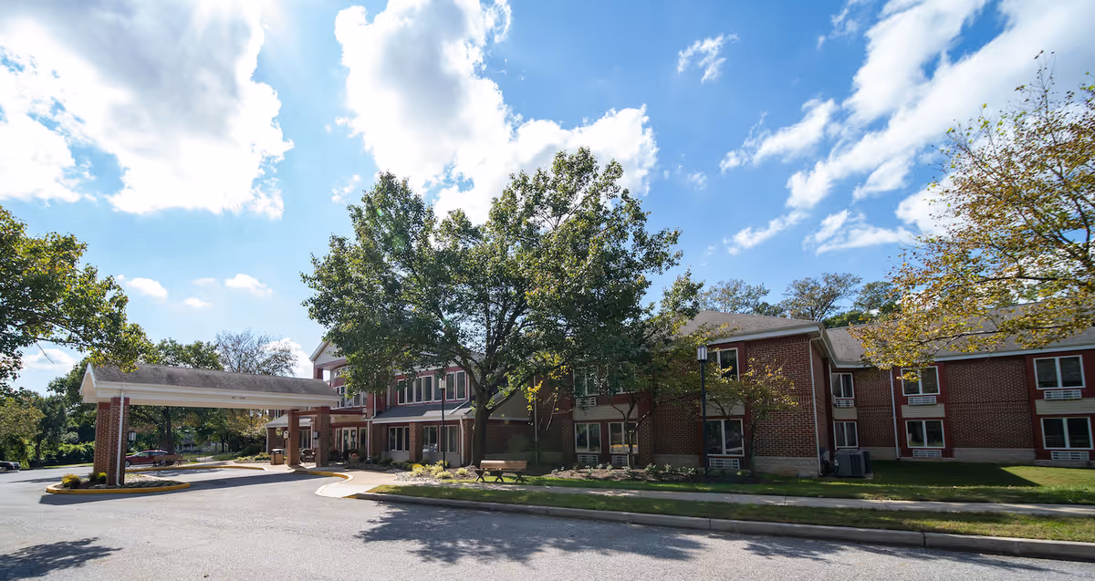 Exterior view of a two-story brick senior living facility building with a covered entrance driveway, surrounded by trees and a clear blue sky with some clouds.