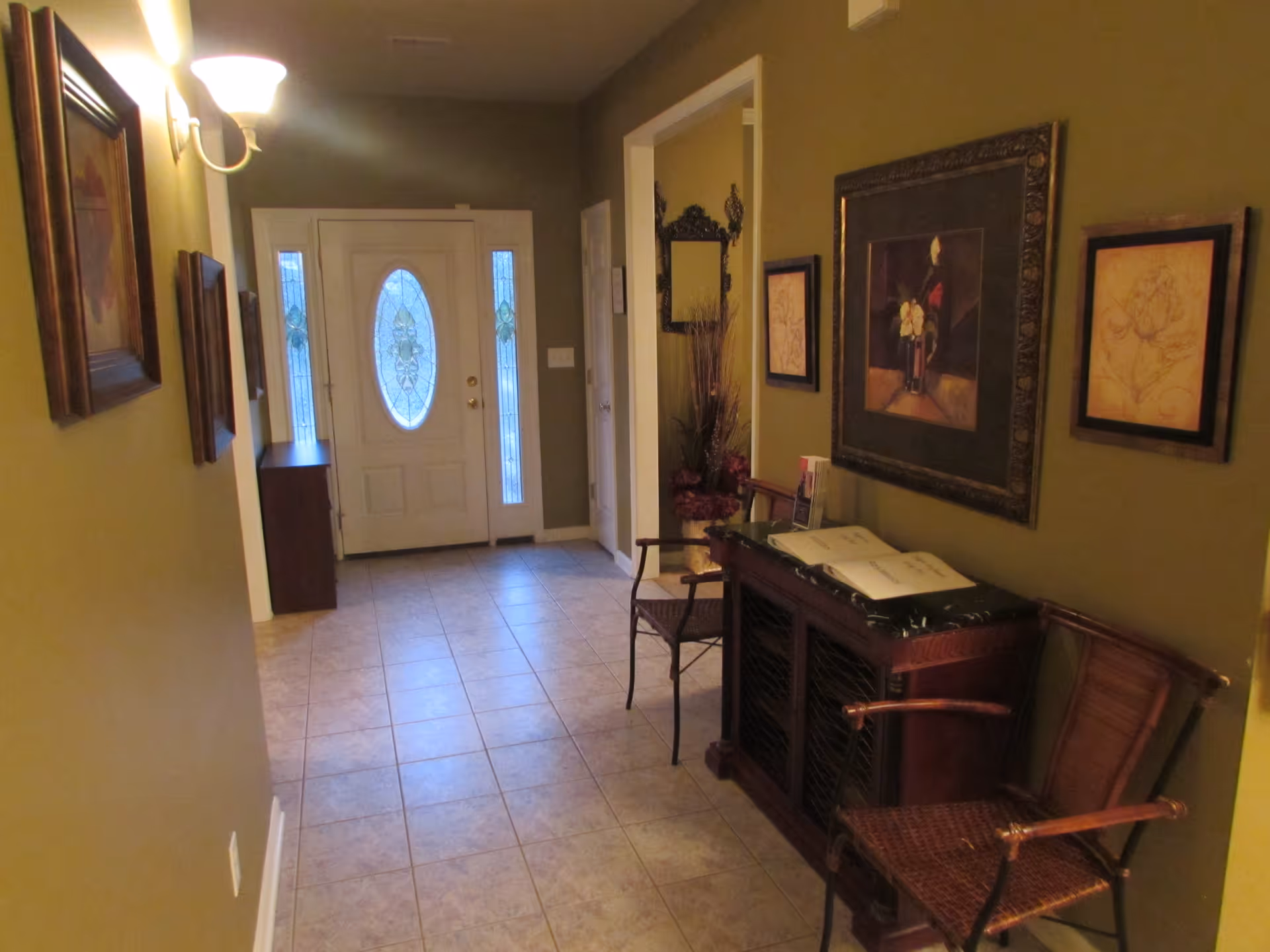 A hallway with tiled floor leading to a white front door with decorative glass panels. The walls are painted olive green and decorated with framed artwork. There is a wooden console table with a book on top and two wicker chairs on either side. A wall-mounted light fixture is visible on the left wall.