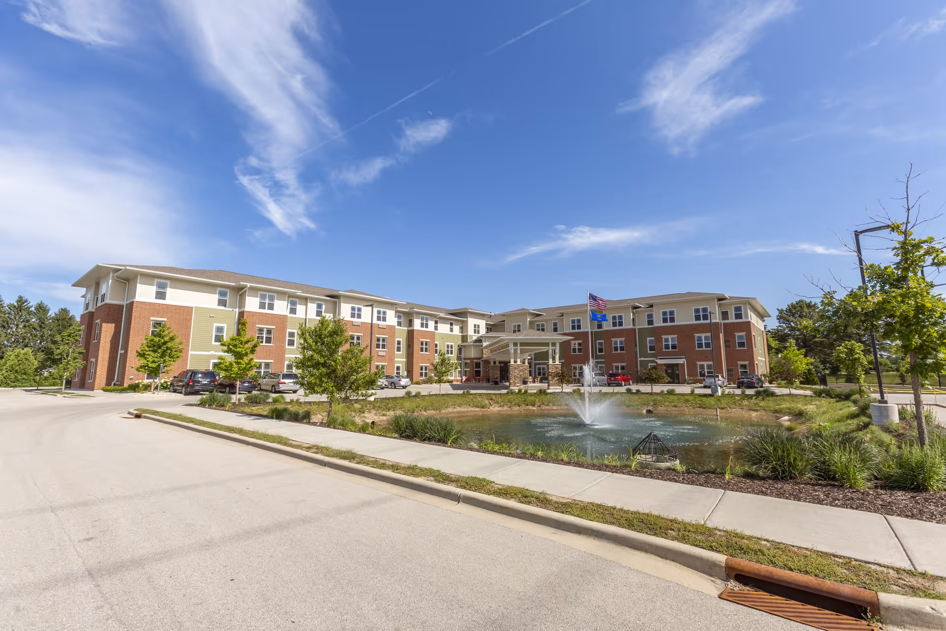 Exterior view of a three-story senior living facility building with a brick and beige facade, surrounded by a parking area, trees, and a small pond with a fountain in front. The sky is clear with some clouds.