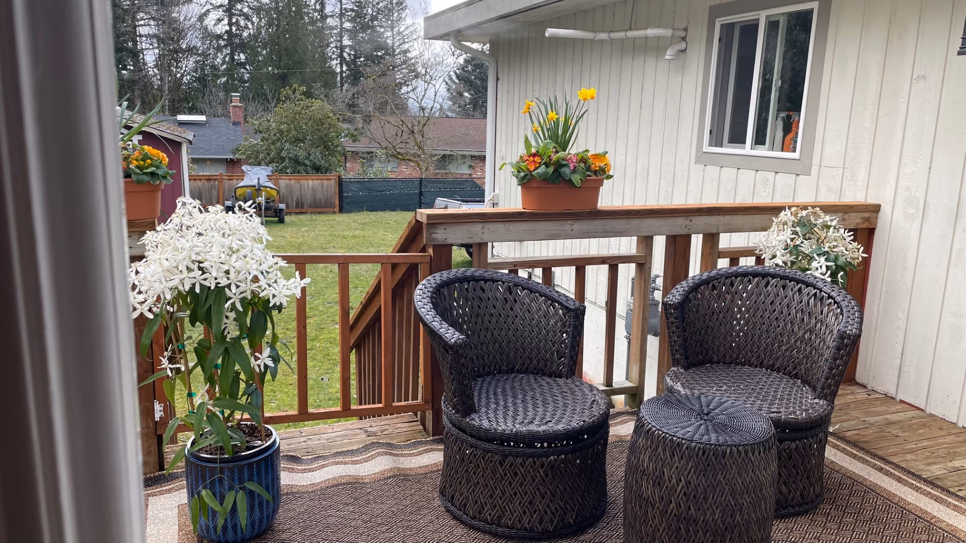 A small wooden deck attached to a light-colored building with two dark wicker chairs and a matching round wicker table. The deck has a brown patterned outdoor rug and several potted plants with white and yellow flowers. In the background, there is a grassy yard with trees and neighboring houses.