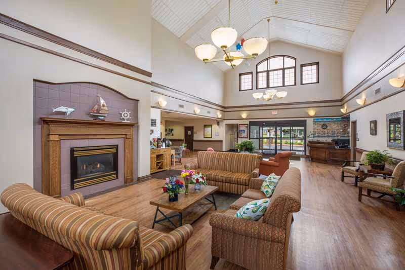 Spacious senior living facility common area with high vaulted ceilings, large windows, and multiple seating arrangements including striped and patterned sofas around a wooden coffee table with flower arrangements. A fireplace with nautical decorations is on the left wall, and a reception desk is visible in the background near the entrance doors.