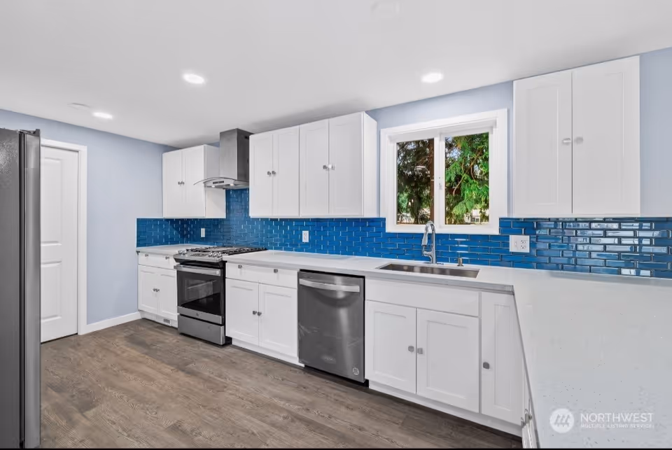 Modern kitchen with white cabinets, blue tiled backsplash, stainless steel appliances including a refrigerator, stove, and dishwasher, a window above the sink, and wood flooring.
