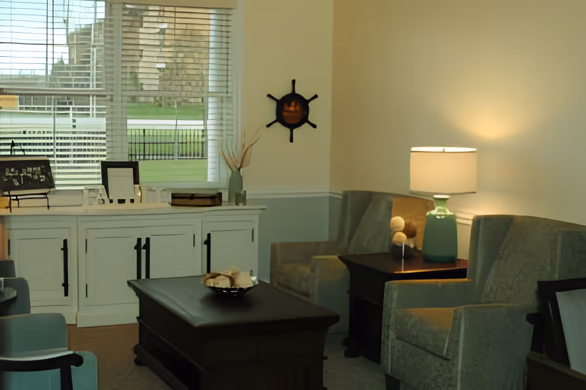 A cozy living room area with two upholstered armchairs, a wooden coffee table with a decorative bowl, a side table with a green lamp, and a white cabinet under a window with blinds. A nautical-themed wall decoration is mounted on the wall.