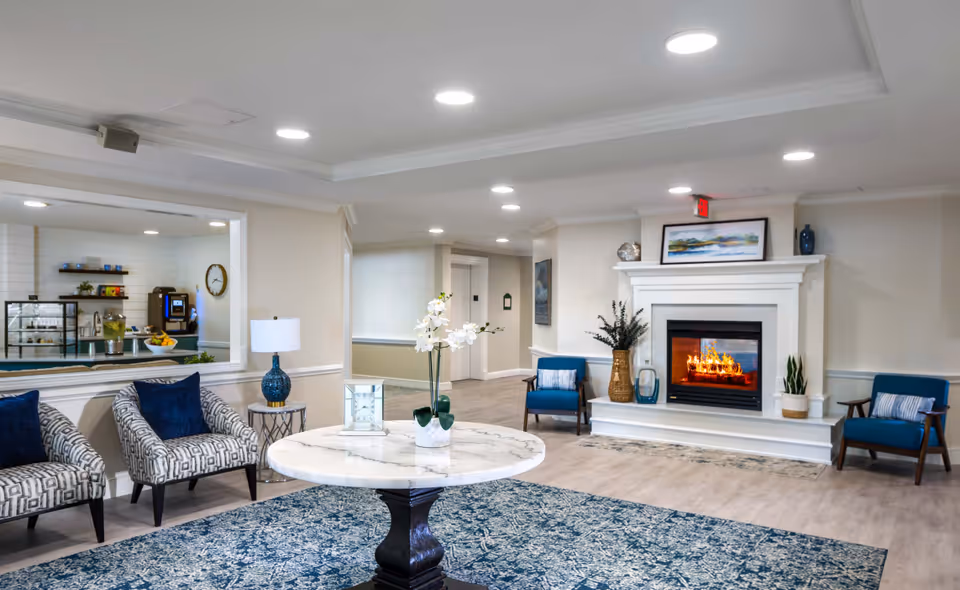 Bright senior living lounge with a fireplace, upholstered chairs, a round marble table, and a coffee bar visible through an interior window.