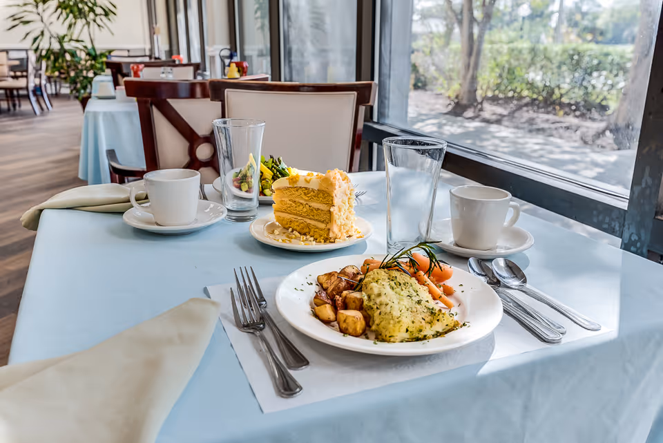 A dining table set with a meal including an herb-crusted fish fillet, roasted potatoes, baby carrots, a slice of layered cake, two cups of coffee, and two glasses of water. The table is covered with a light blue tablecloth and is next to a large window with a view of greenery outside. The background shows more tables and chairs in a dining room setting.