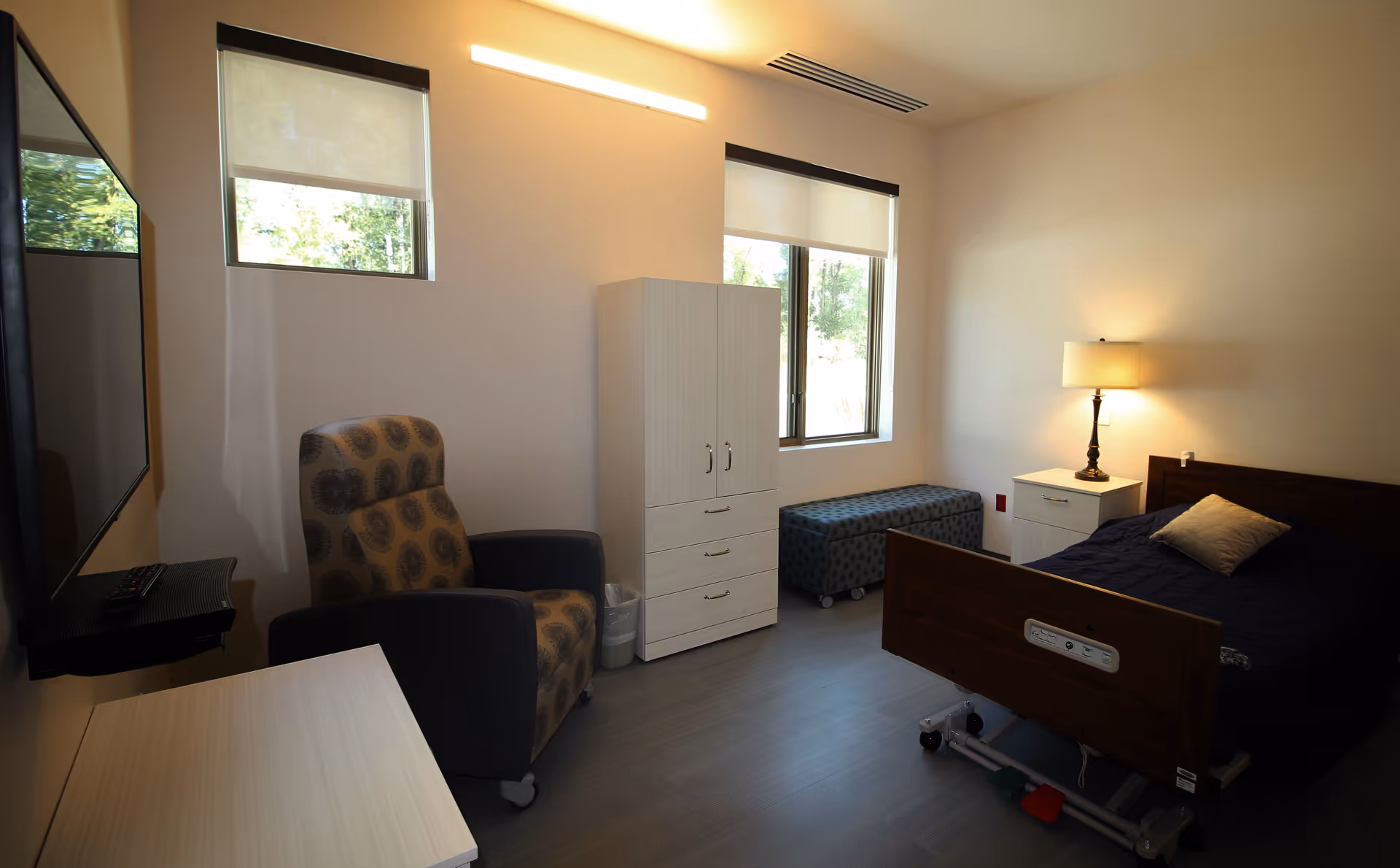 A senior care private bedroom with a hospital-style bed, armchair, wardrobe, wall-mounted TV, and windows.