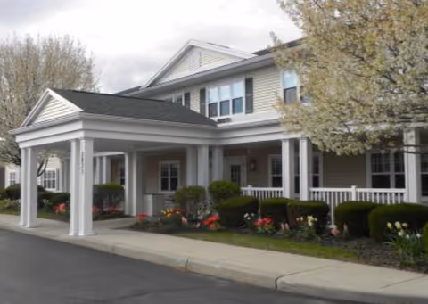 Exterior view of a senior living facility building with a covered entrance supported by white columns. The building has beige siding, white trim, and multiple windows. There are flowering trees and landscaped bushes with colorful flowers along the front walkway.