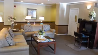 A cozy common lounge with sofas, a coffee table with magazines and flowers, table lamps, and a TV cabinet.