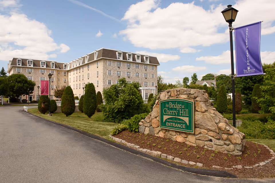 Exterior view of The Bridge at Cherry Hill assisted living community building with a stone sign at the entrance and landscaped greenery under a partly cloudy sky.
