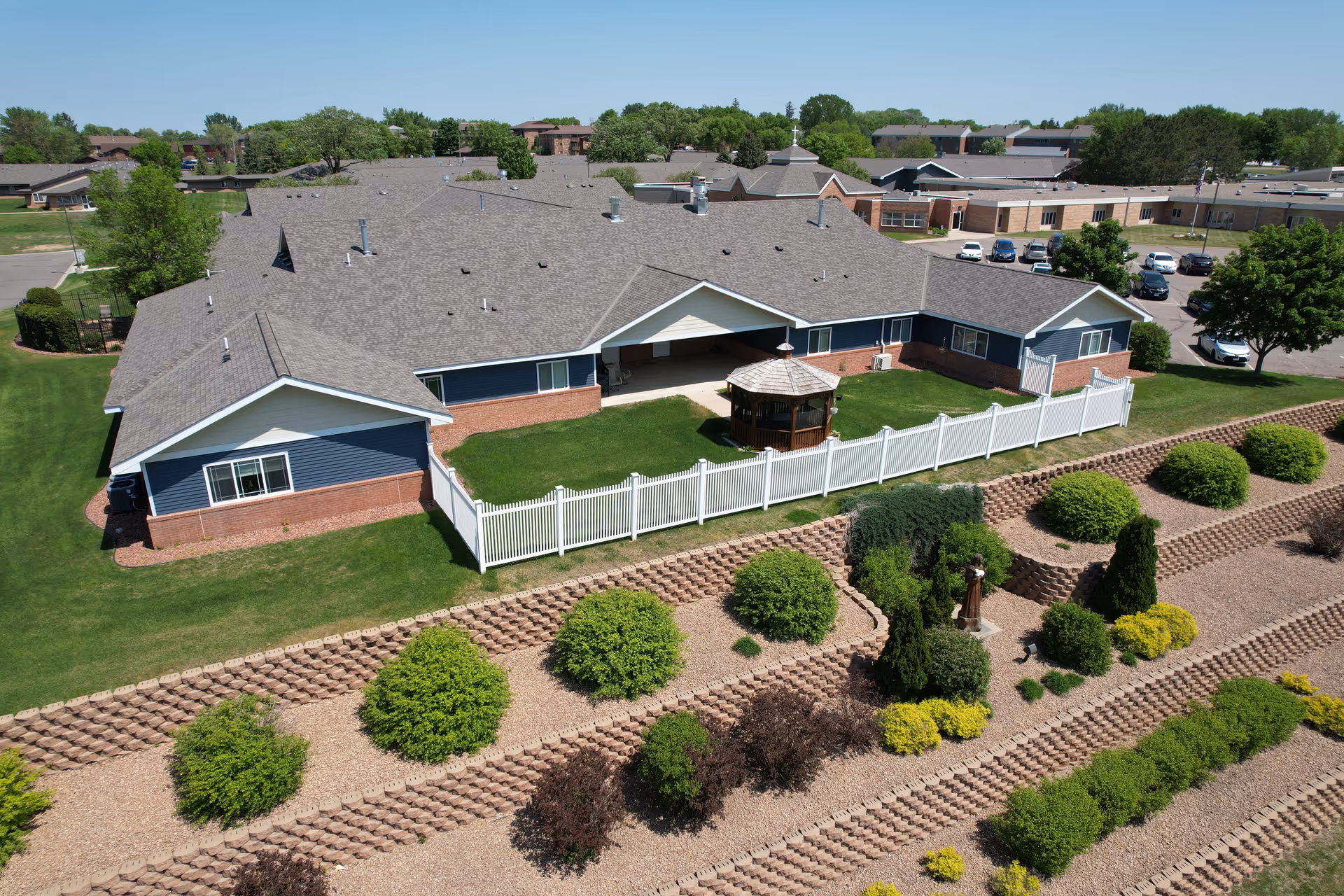 Aerial view of a single-story senior living building with a white fenced courtyard, central gazebo, and terraced landscaping.