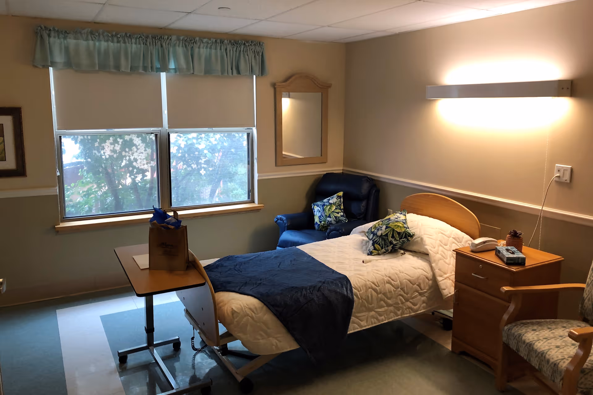 A well-lit bedroom in a senior living facility with a single bed covered in a white quilt and a dark blue throw blanket. There are two decorative pillows on the bed. Next to the bed is a wooden nightstand with a telephone and a box of tissues. A blue armchair with a floral pillow is placed in the corner near a window with closed blinds and a green valance. A wooden-framed mirror hangs on the wall above the armchair. A small overbed table with a brown paper bag is positioned next to the bed. The room has light-colored walls and a patterned floor.