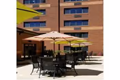 Outdoor patio with multiple tables, chairs, and umbrellas beside a multi-story brick building.