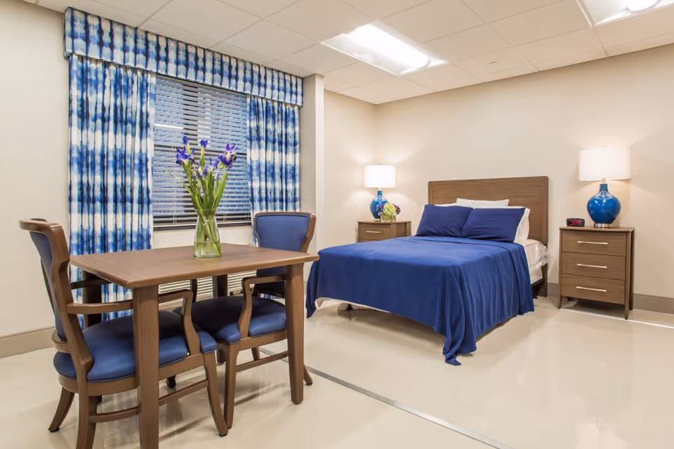 A bright and clean bedroom in a senior living facility featuring a bed with blue bedding, two wooden nightstands with blue lamps, a wooden table with four blue cushioned chairs, and a window with blue and white patterned curtains. A vase with purple flowers is placed on the table.